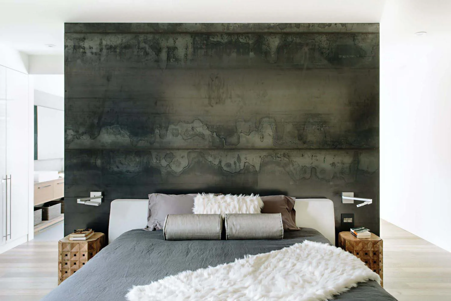 A modern industrial-style bedroom with a concrete textured headboard wall, a grey bed featuring soft pillows and a fur blanke