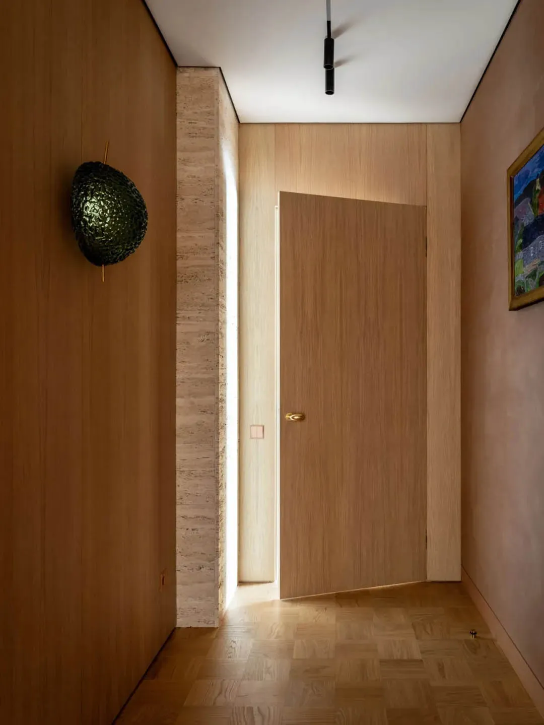 Modern minimalist entryway with wooden flooring and door, featuring a green textured wall sconce and illuminated corridor.