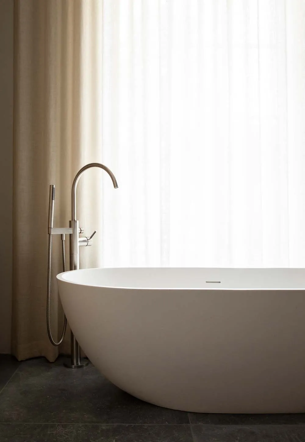 A minimalist bathroom with a large ceramic bathtub, a modern faucet, and a sheer curtain.