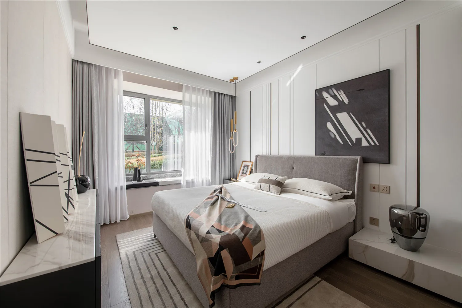 Modern minimalist bedroom featuring a grey upholstered bed with a geometric abstract painting above, white curtains, and a st
