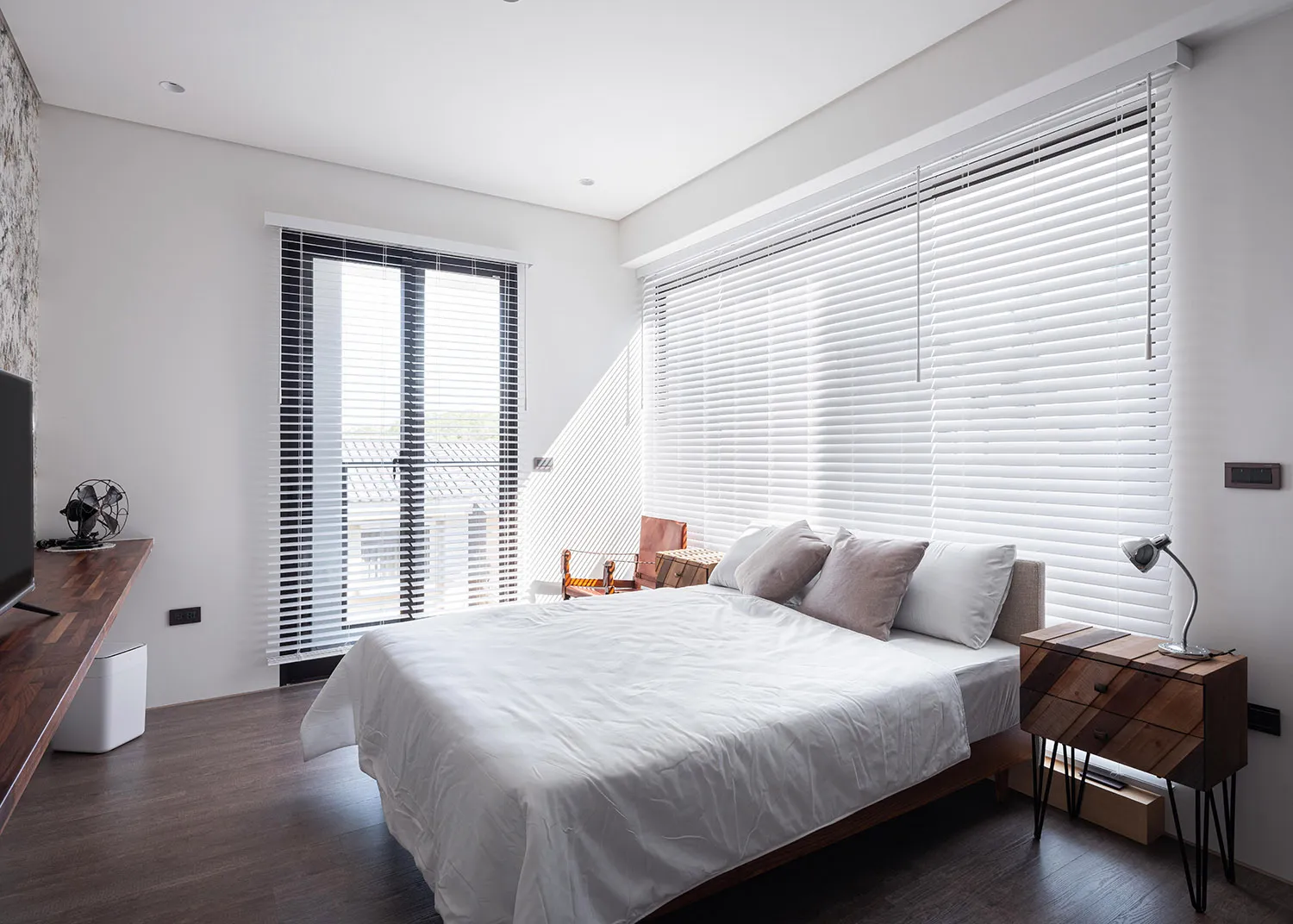 A minimalist bedroom with a large bed, white bedding, and white blinds covering the windows.