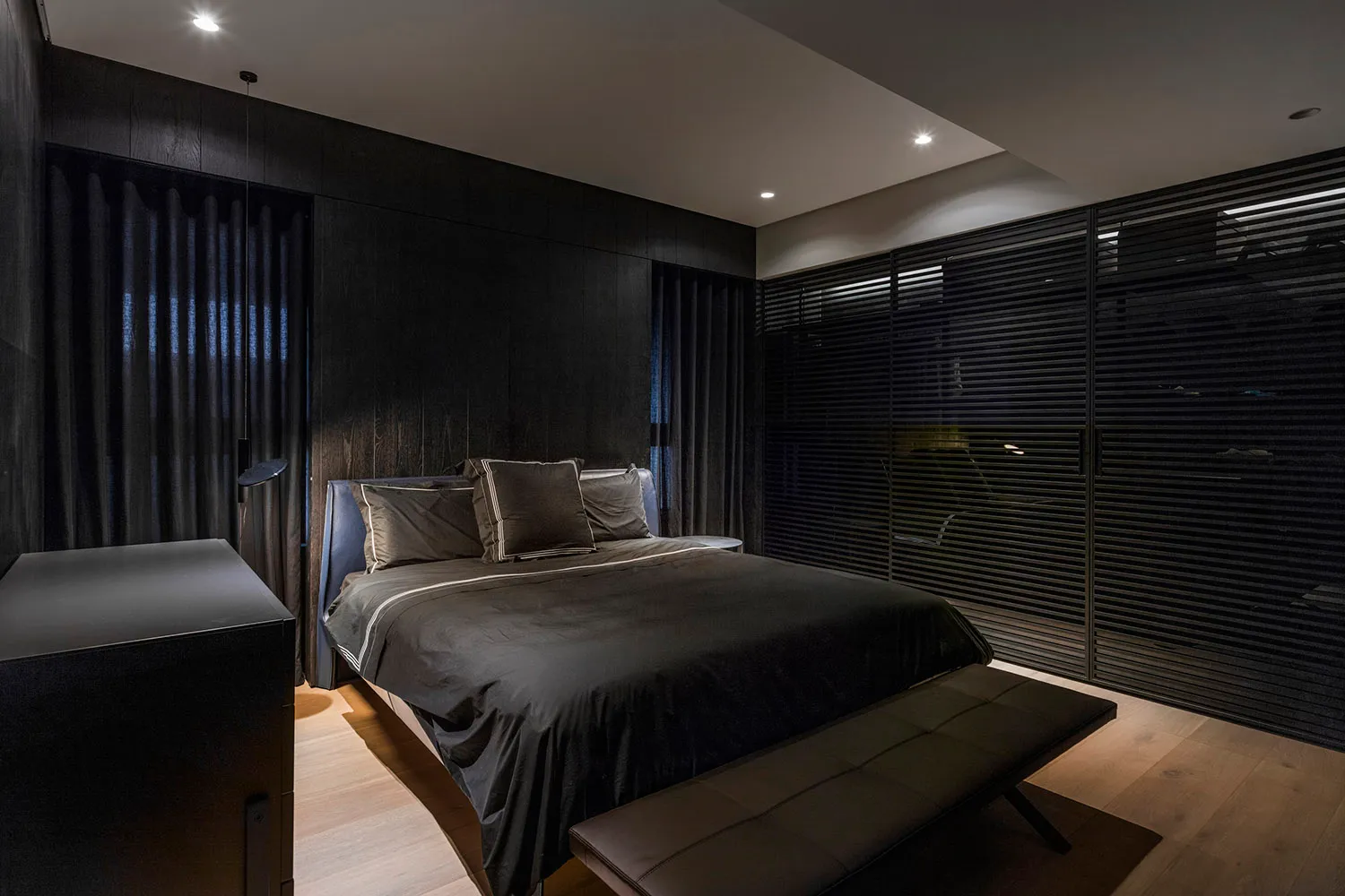 Minimalist bedroom featuring a wood bed with black bedding, a bench, and dark curtains.
