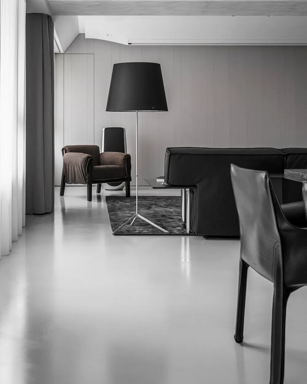 A minimalist living room with concrete flooring, a black sofa, a brown armchair, and a black floor lamp.