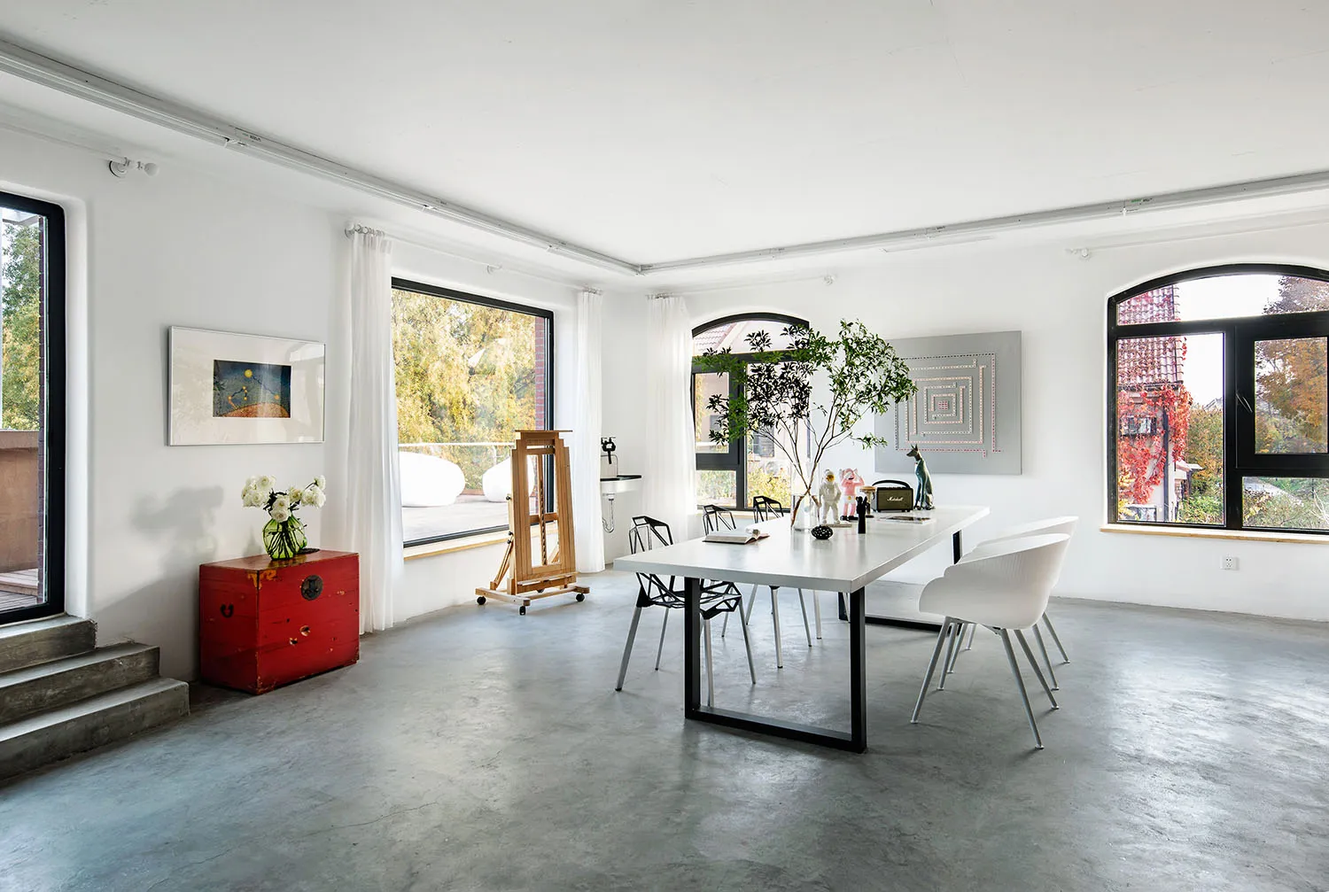 A spacious, modern dining room featuring a white table, white chairs, and a concrete floor with arched windows, artwork, and