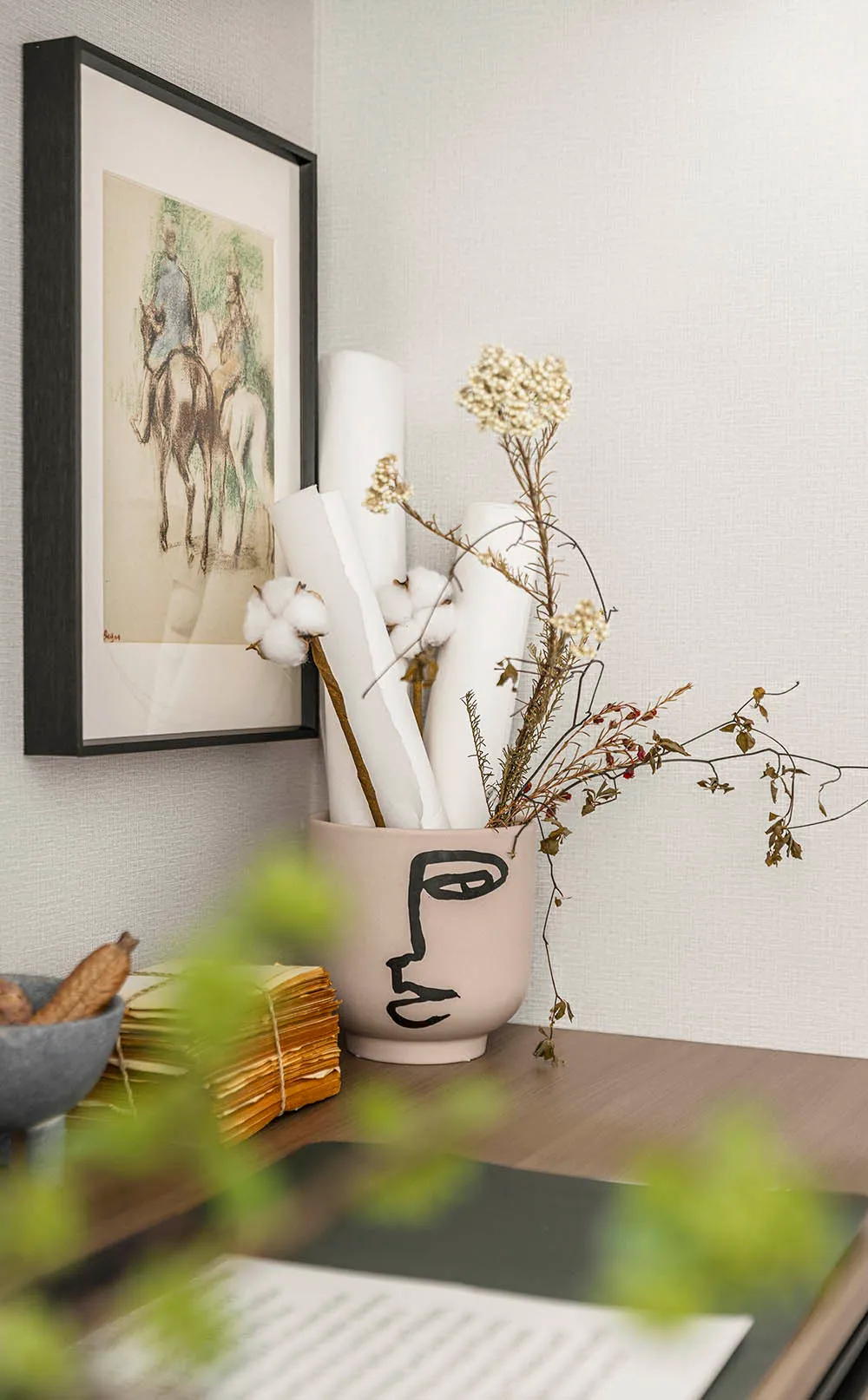 A modern decorative arrangement featuring a ceramic vase with a face design, flowers, and paper rolls on a wooden surface.