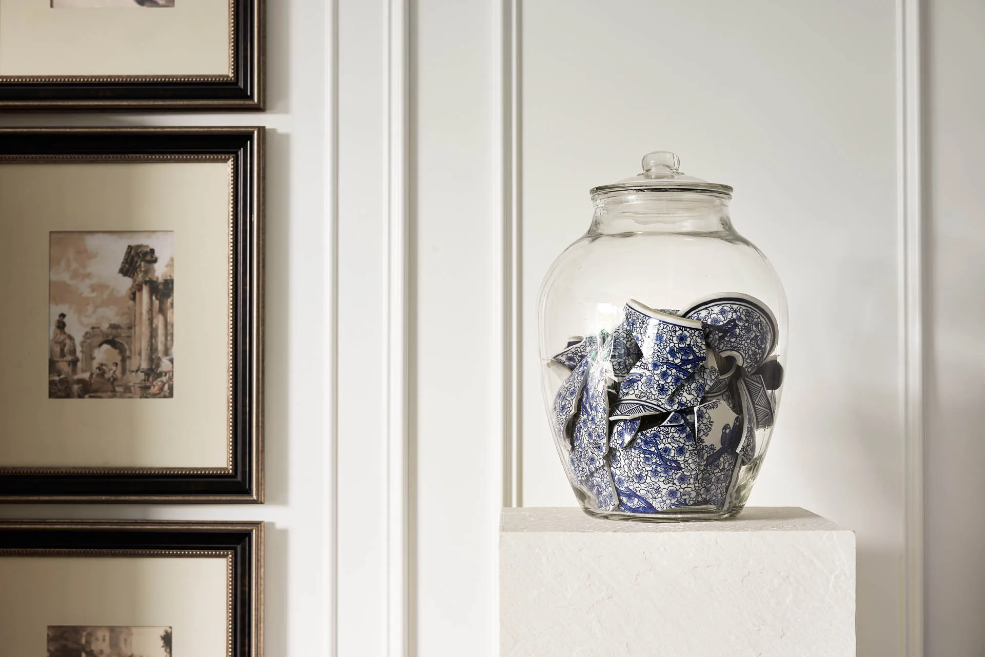 A glass jar containing broken pieces of blue and white ceramic vases is placed on a white pedestal against a white wall adorn