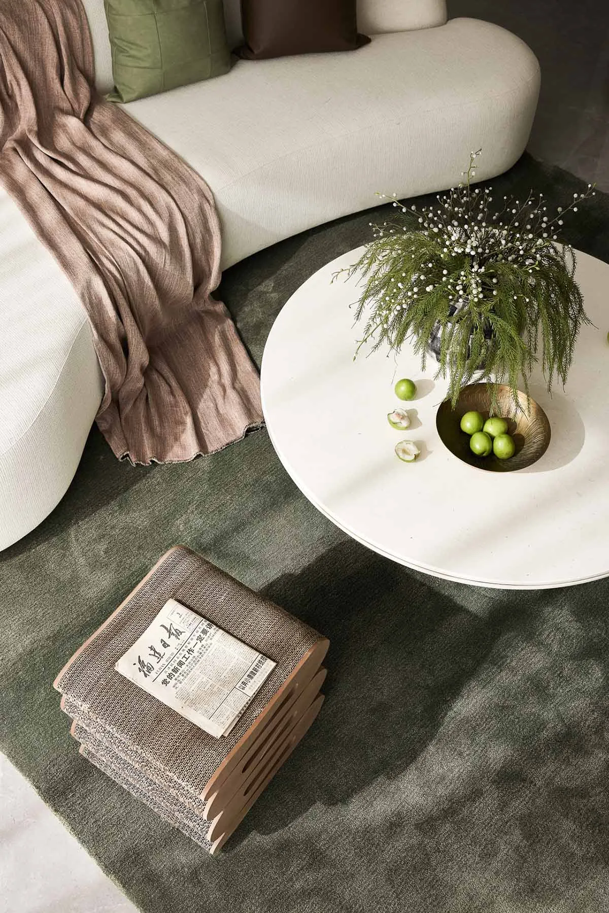A modern living room featuring a white curved sofa, a round white coffee table with greenery and fruits, and a stack of books