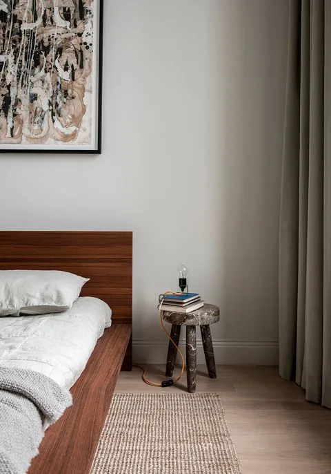 A minimalist bedroom featuring wooden furniture and a hanging light bulb on a stool, with a framed abstract painting above th