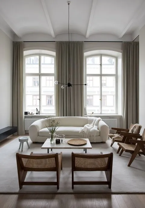 A modern minimalist living room featuring a white cushioned sofa, wicker chairs, and large windows with grey curtains.