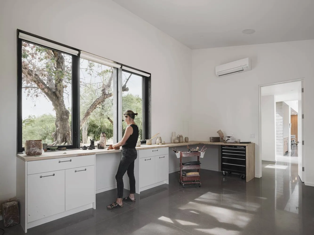 A modern studio workspace featuring a large window, wood counters, metal shelves, and a person working on a piece of pottery.