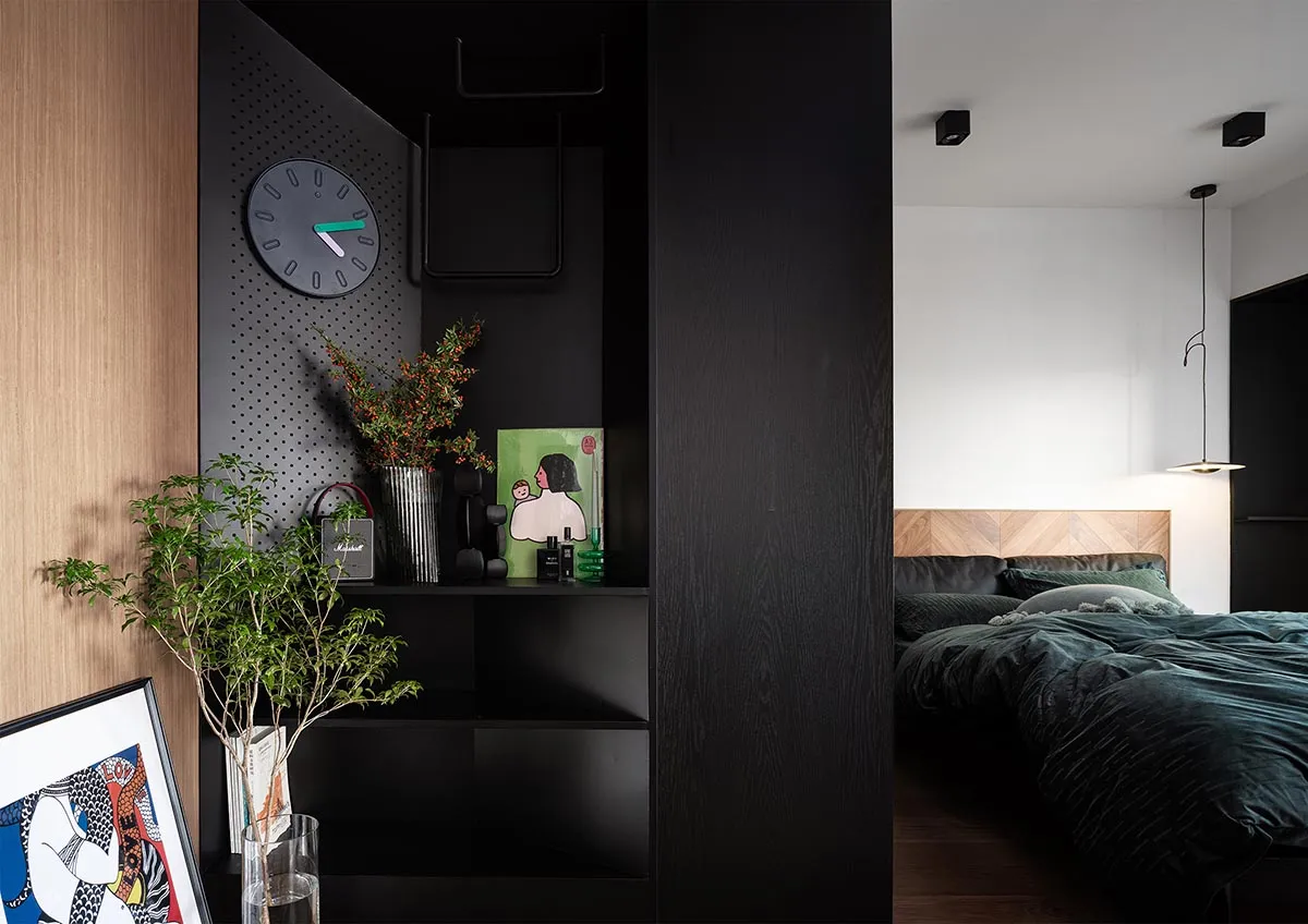 A modern bedroom featuring a dark-colored shelf with a clock, plants, and decor, leading to a bed with velvet bedding and a w