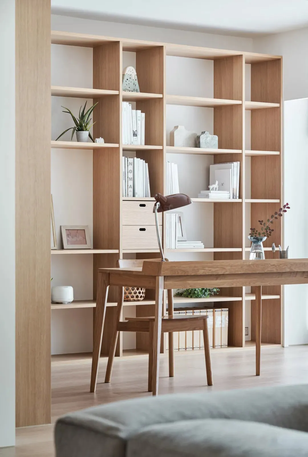 A minimalist home office featuring light wood shelves, a wooden desk, and a grey sofa.