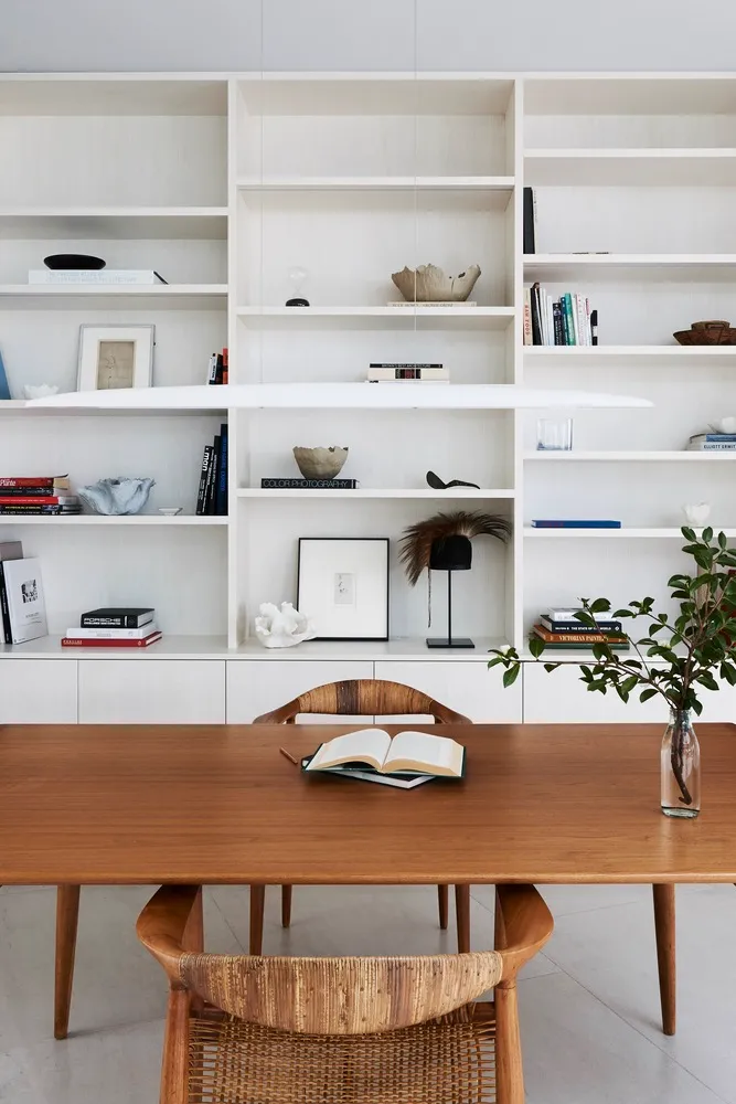 A modern home office with a wooden table, leather chairs, and white bookshelves filled with books and decorative items.