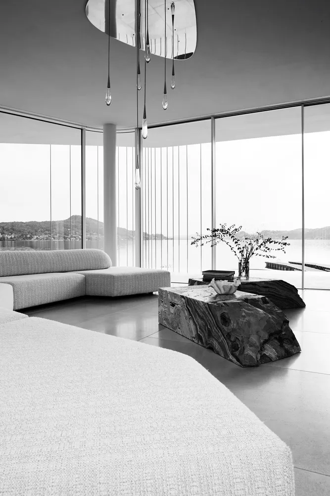Modern minimalist living room with concrete floor, sofa, and coffee table, large window overlooking water.
