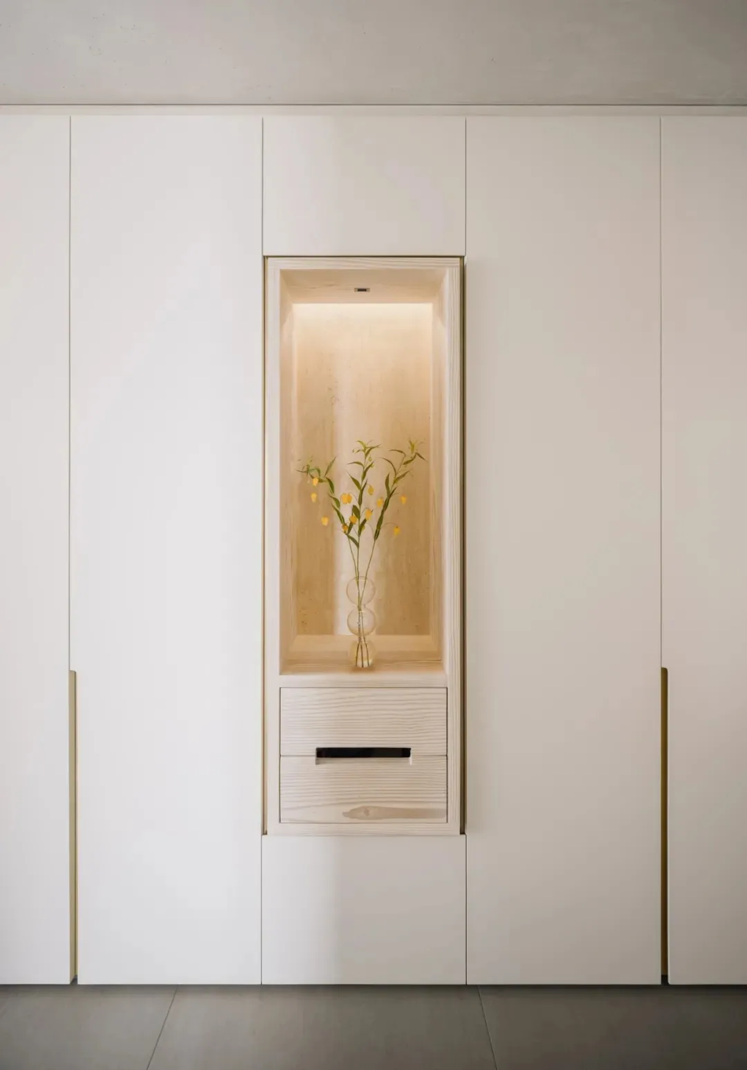 A minimalist interior wall niche with a wooden frame, illuminated light, and a vase of flowers.