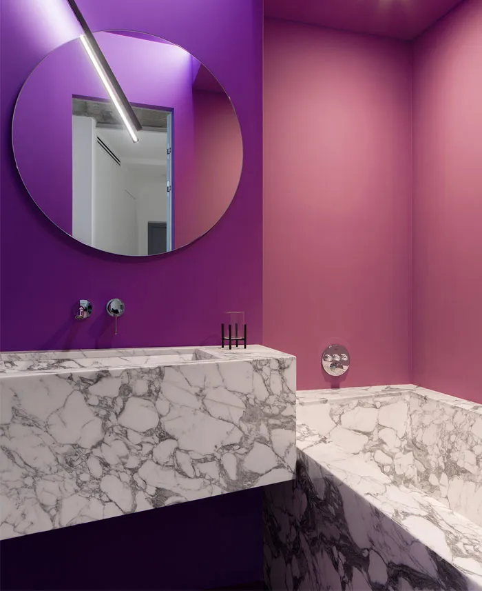 A modern interior bathroom with purple and pink walls, marble sink, and a large round mirror.