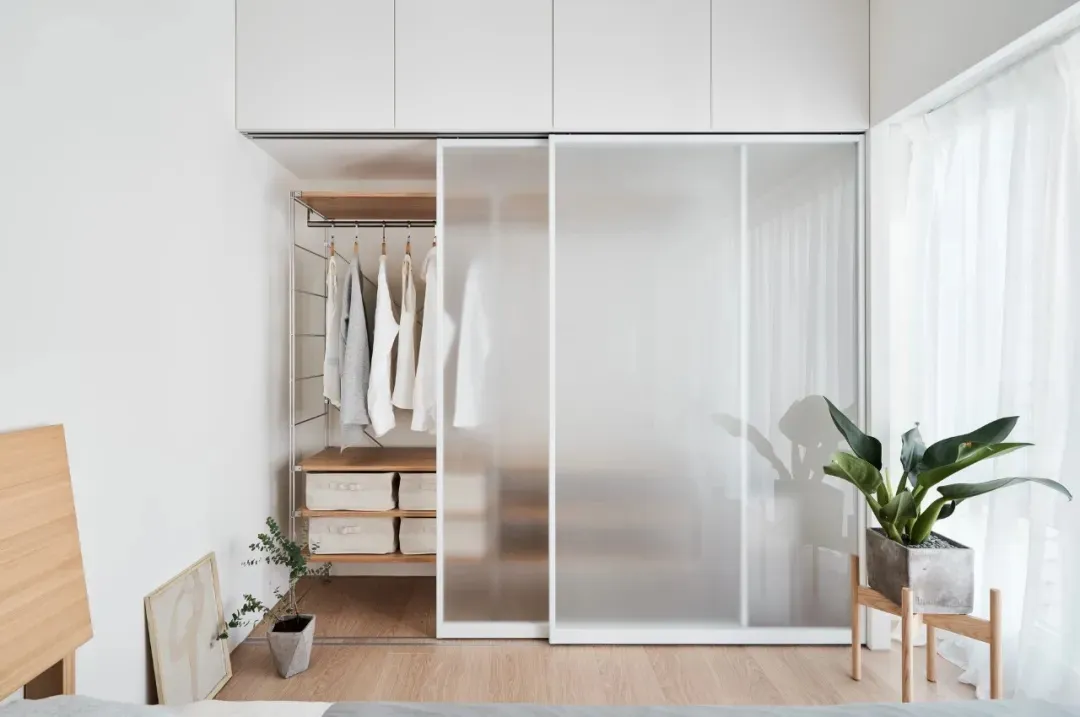 A minimalist bedroom with a glass sliding door, white furniture, and light wood flooring.