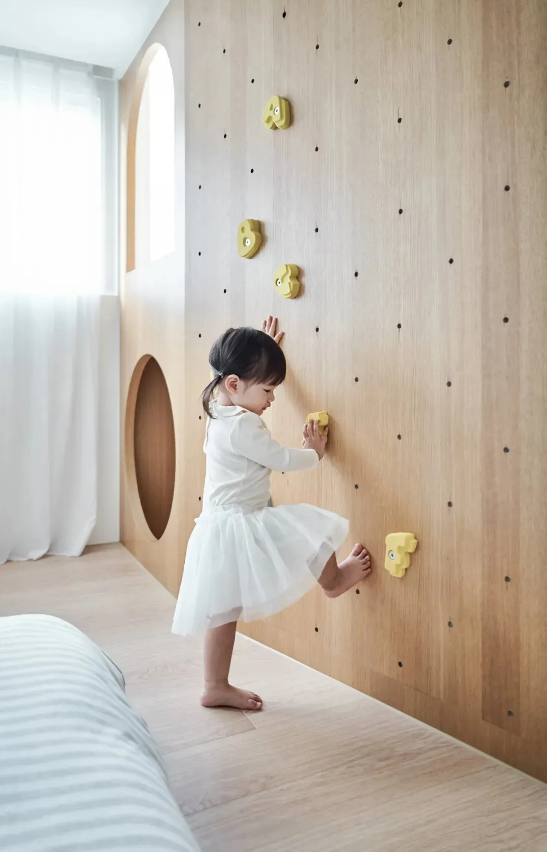 一位幼兒在現代兒童遊戲室內攀爬木質牆面,牆上裝有黃色攀爬握把。