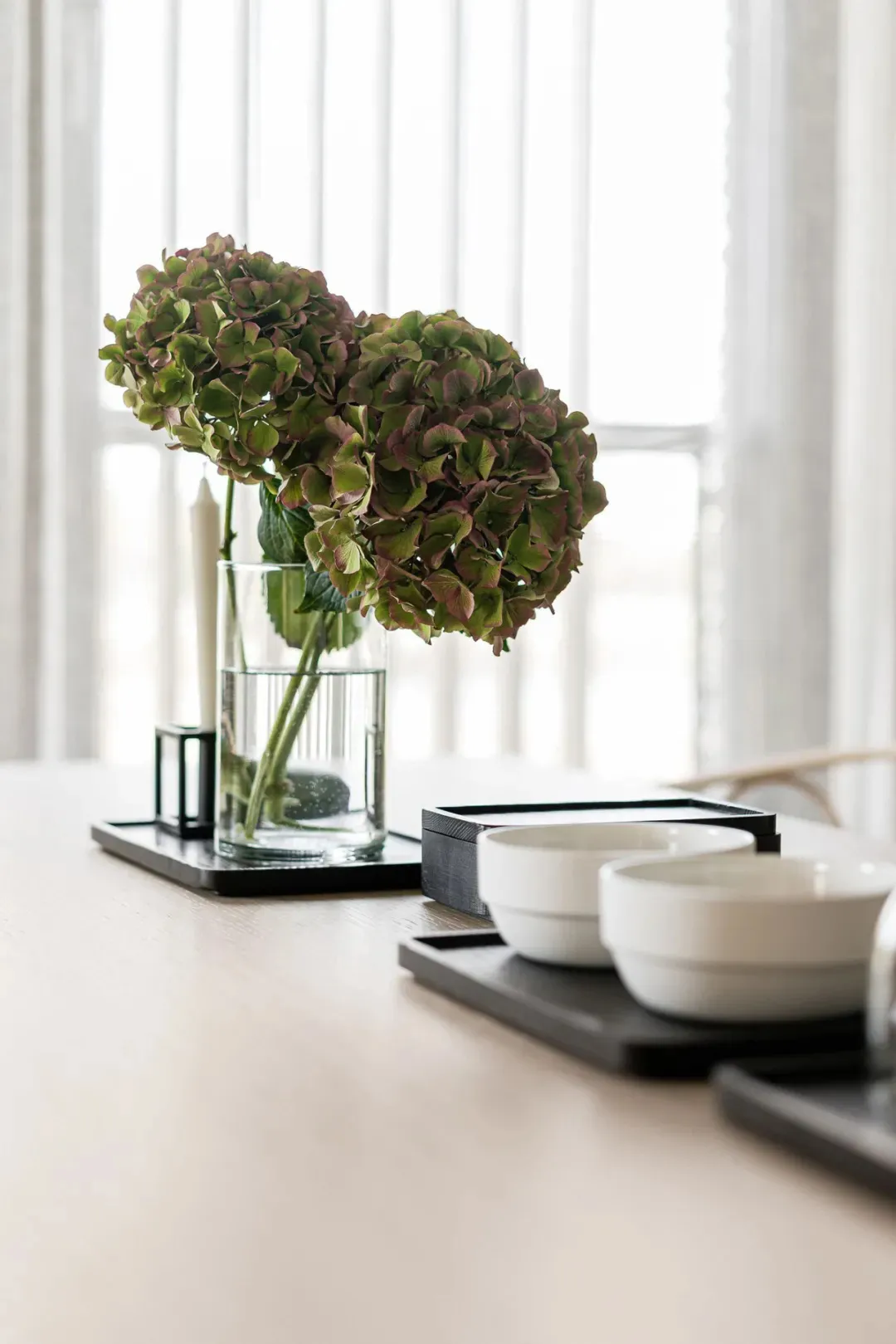 A minimalistic table setting with a clear glass vase containing hydrangea flowers and bowls on a tray.