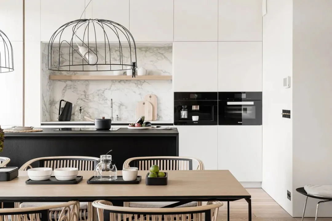 A modern, minimalist kitchen with wooden dining table, marble backsplash, and metal pendant lights.
