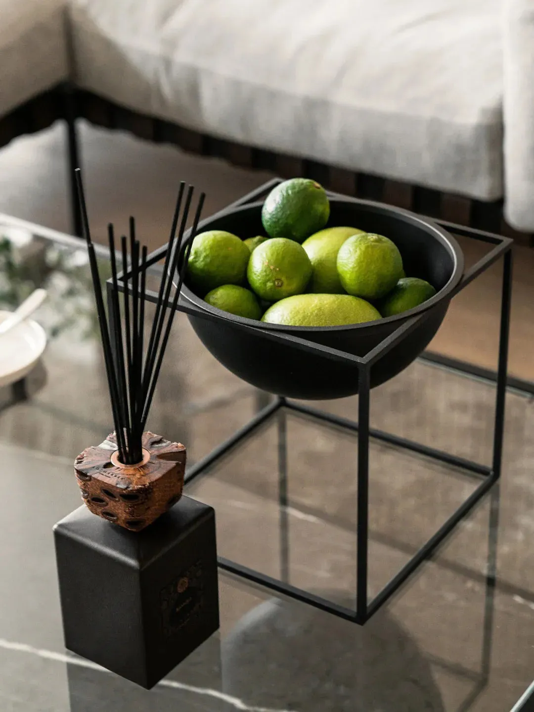 A glass table with a modern black metal frame holds a bowl filled with green limes and a brown ceramic vase filled with black