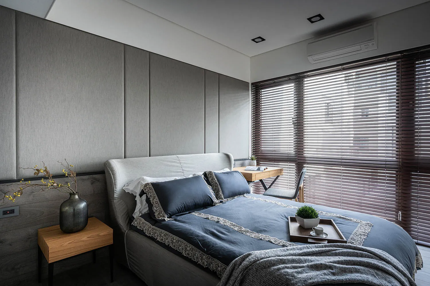 A modern minimalist bedroom featuring grey upholstered walls, wooden furniture, and a small wooden desk with a plant and cup.