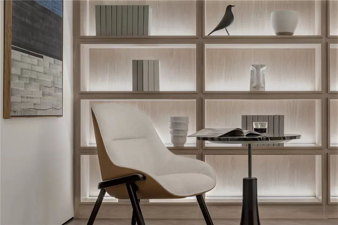 Minimalist living room with a modern white armchair, a black side table, a wall-mounted bookshelf with books, a white vase, a