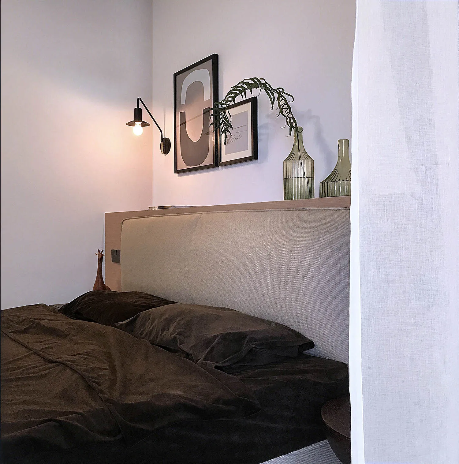 A cozy, minimalistic bedroom featuring dark textiles, a structured headboard, and a wall-mounted light fixture.