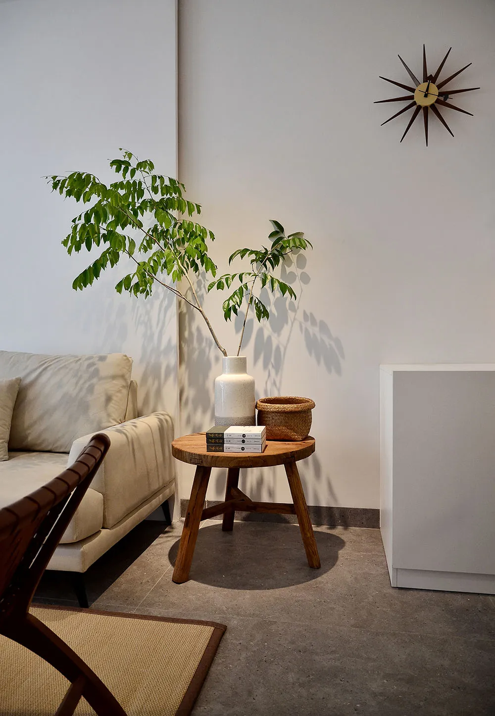 Minimalist living room featuring a wooden stool with a vase, a small plant, a woven basket, and a stack of books next to a wh