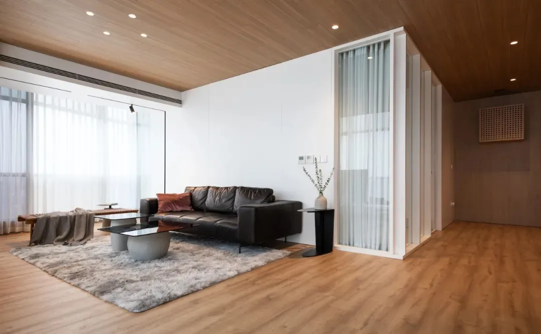 A contemporary living room with a black leather sofa, wooden flooring, and large floor-to-ceiling windows.