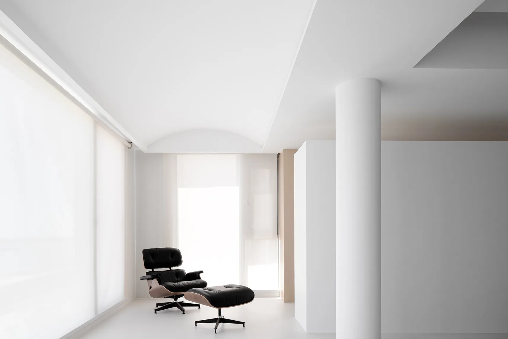 A minimalist office space featuring a white interior with a black chair and ottoman, large windows, and a sleek design.