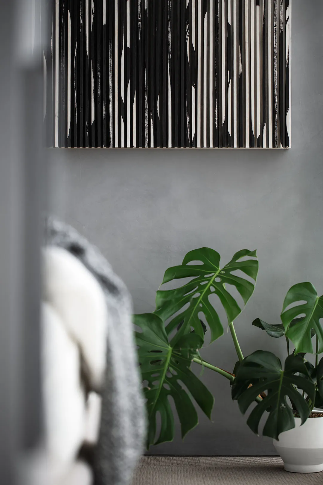 A monstera plant in a concrete planter with a canvas artwork featuring vertical black and white lines hanging on a matte gray