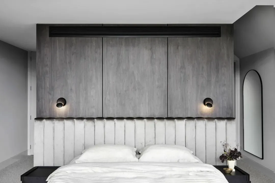 A minimalist bedroom with a wooden headboard and white bedding, featuring grey walls and a mirrored accent wall.