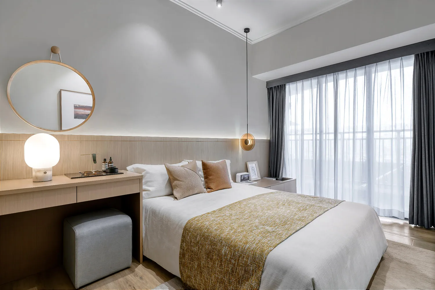 A modern bedroom featuring a white bed with gold accents, a wooden desk, and a large window with sheer curtains.