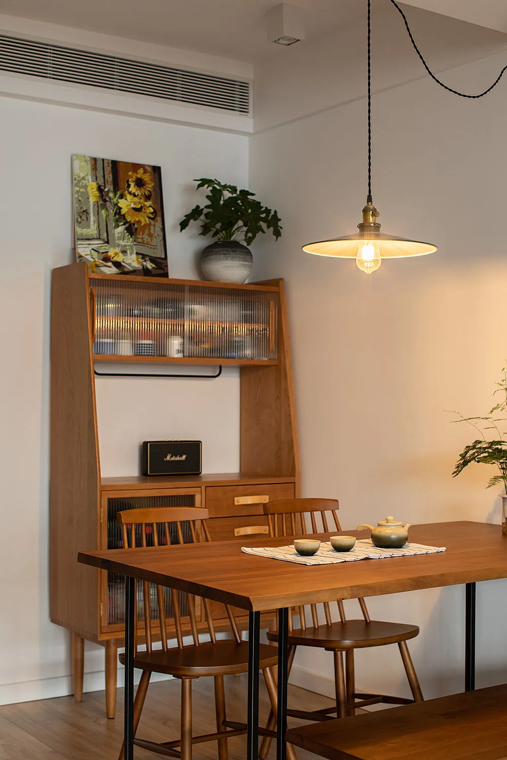 Modern kitchen with a table, chairs, and shelves made of teak wood, featuring a potted plant and a hanging light fixture.