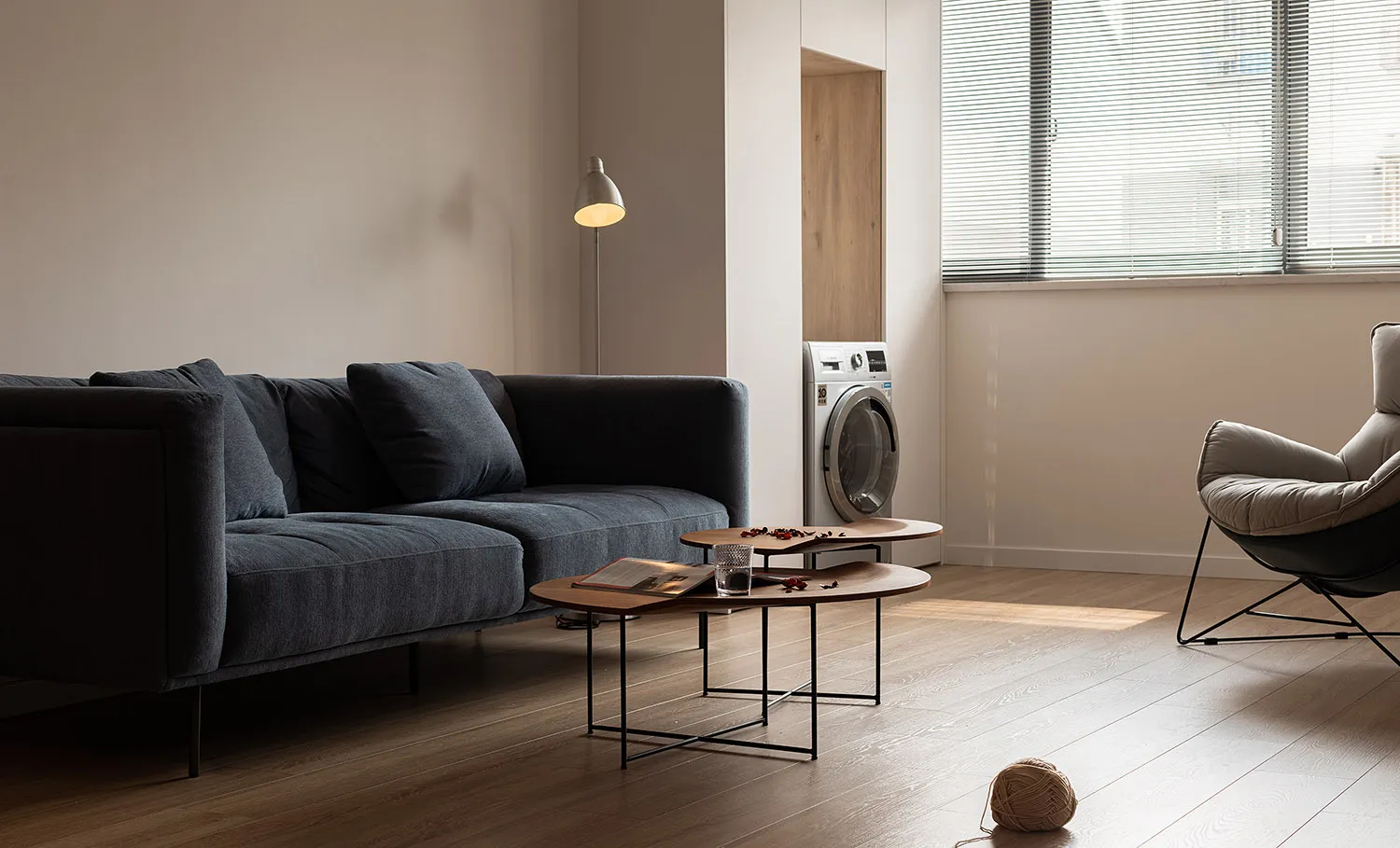A modern living room featuring a wooden floor, a dark gray sectional sofa, a coffee table, a standing lamp, and a window with