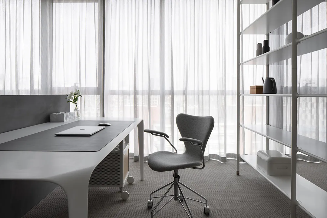 A modern office space with a minimalist design featuring a white desk, gray rolling chair, and white shelves.