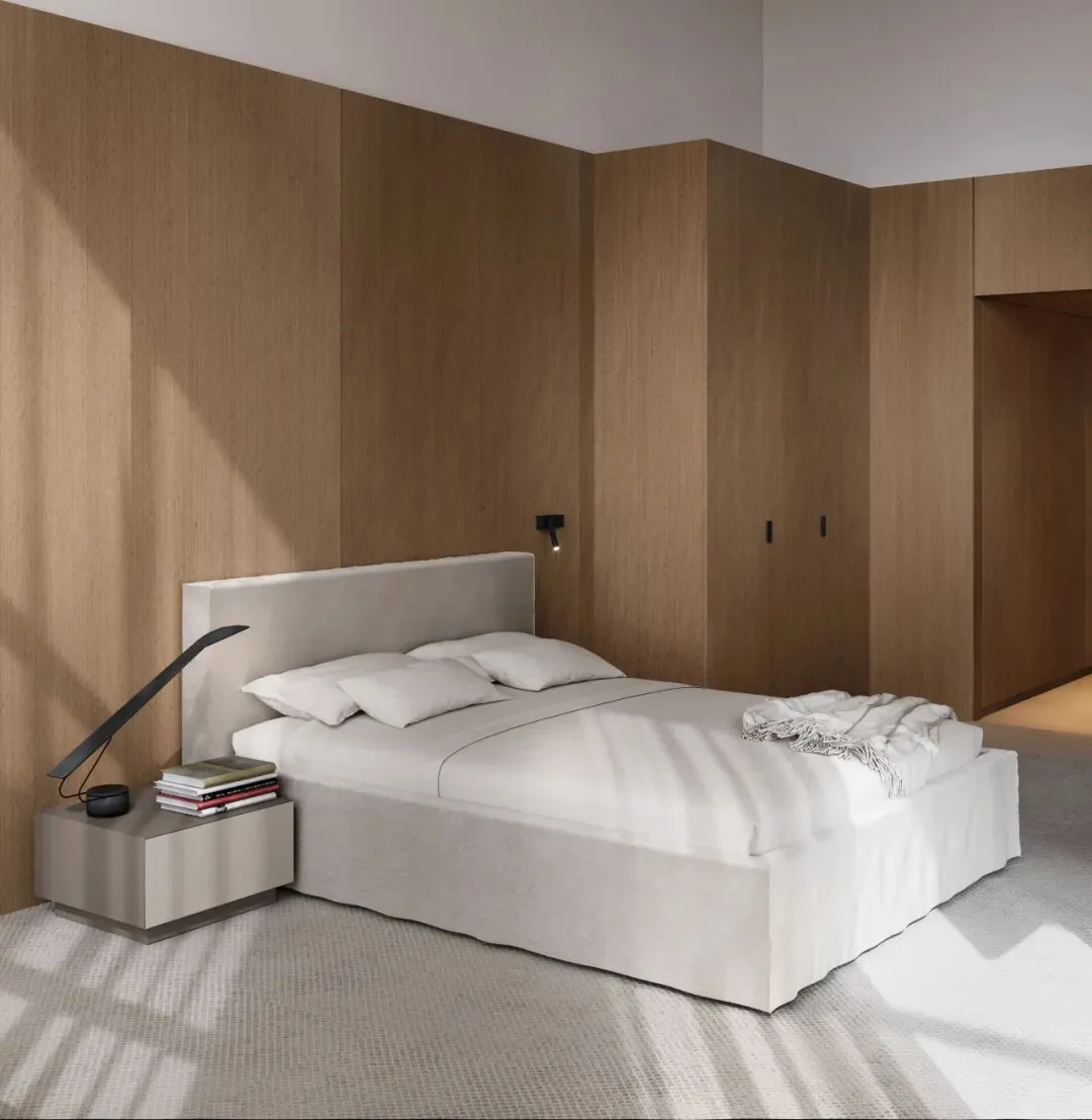 A minimalist bedroom with a white bed sheet, a headboard, and a nightstand with a lamp and books.