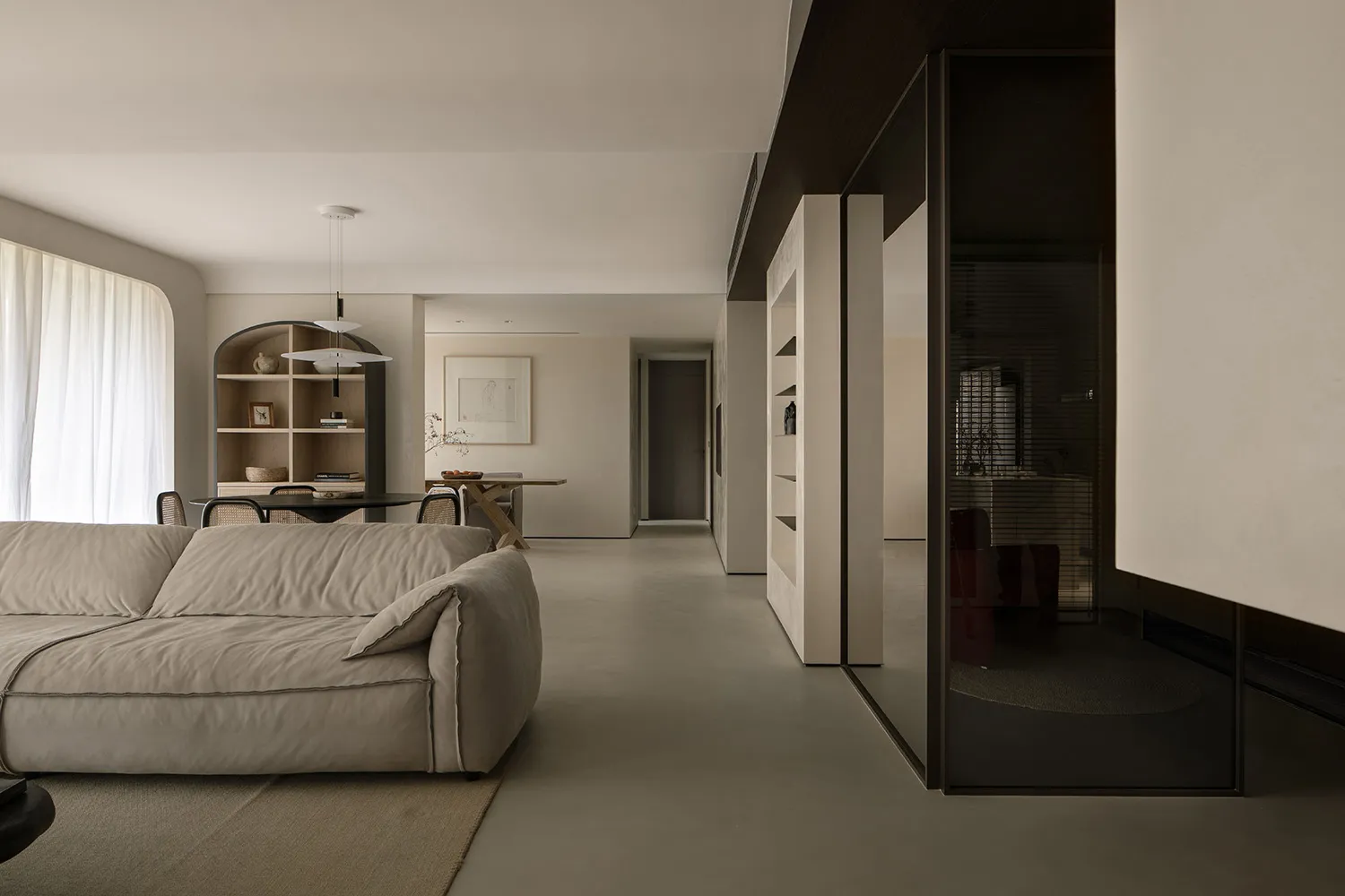 A minimalist living room with neutral-colored furniture and walls, featuring a lounge sofa, dining table, shelves, and a glas