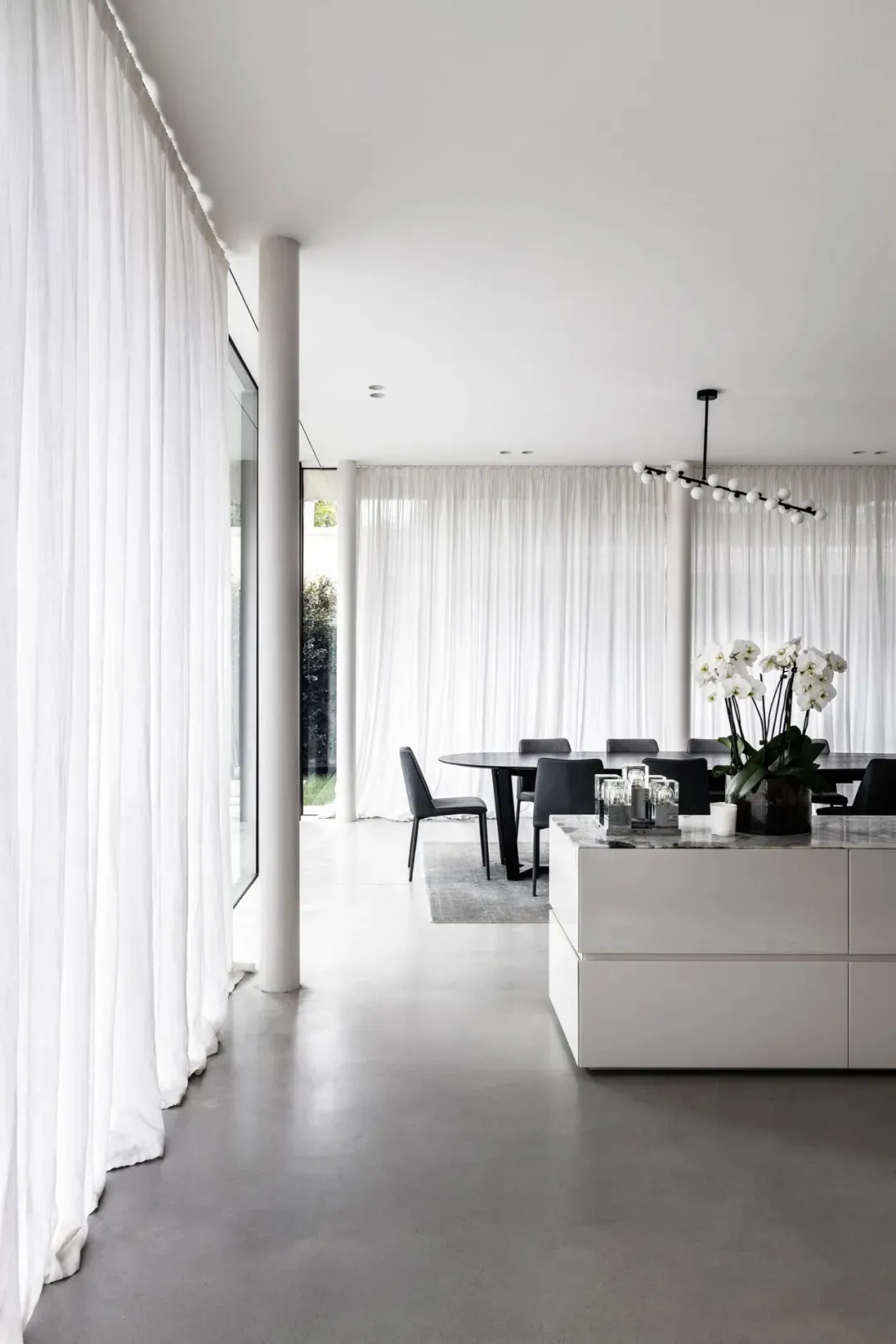 A modern minimalist dining room with white curtains, black furniture, concrete floors, a glass chandelier, and a marble dinin