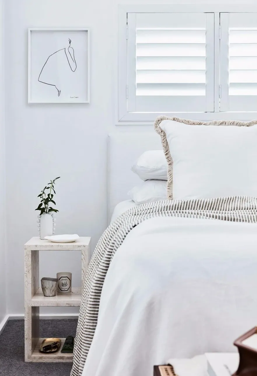 Minimalist bedroom with white walls, white bedding, and a small nightstand with greenery and candles.