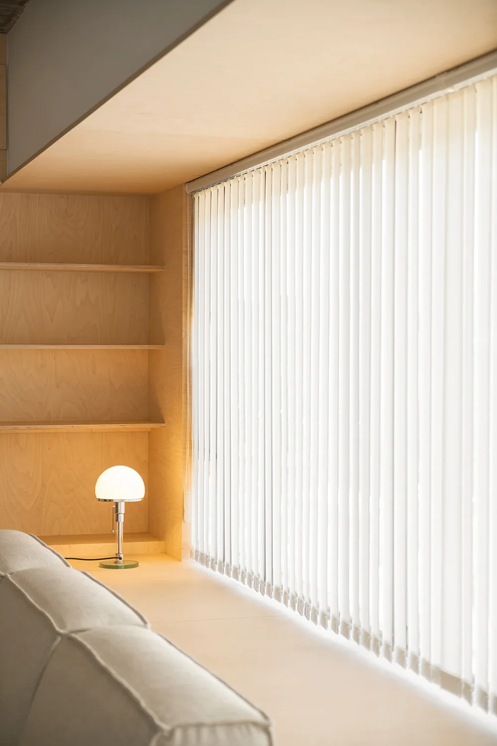 Beige couch against a wall with white vertical blinds and wooden shelves.