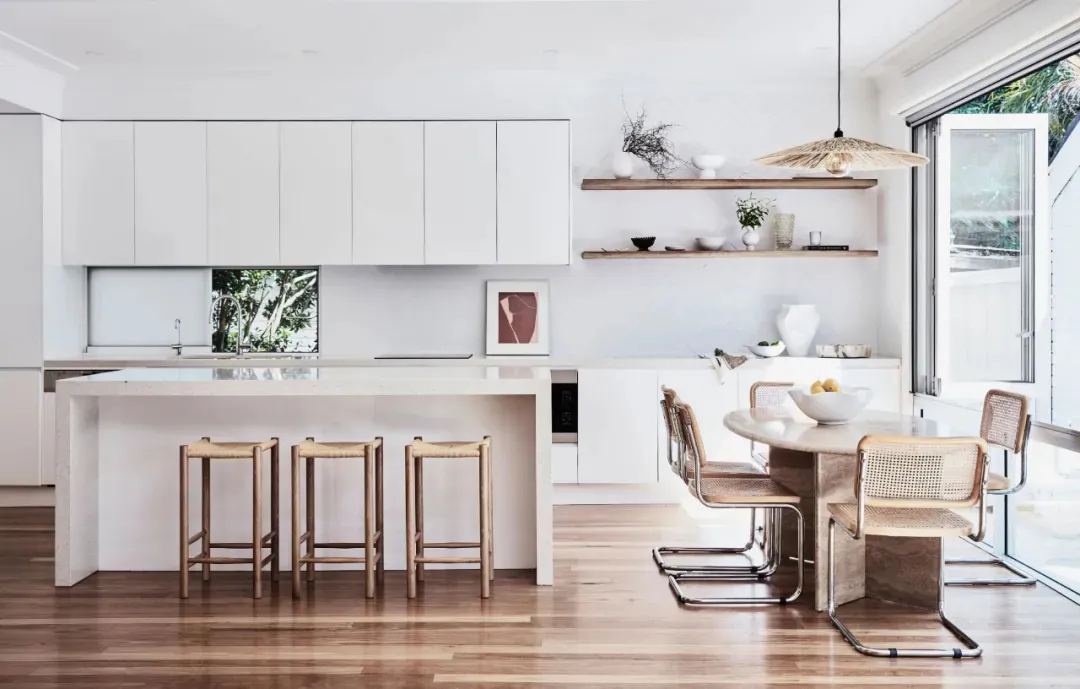 A modern kitchen featuring sleek, white cabinets, wooden stools, and an open shelving unit.