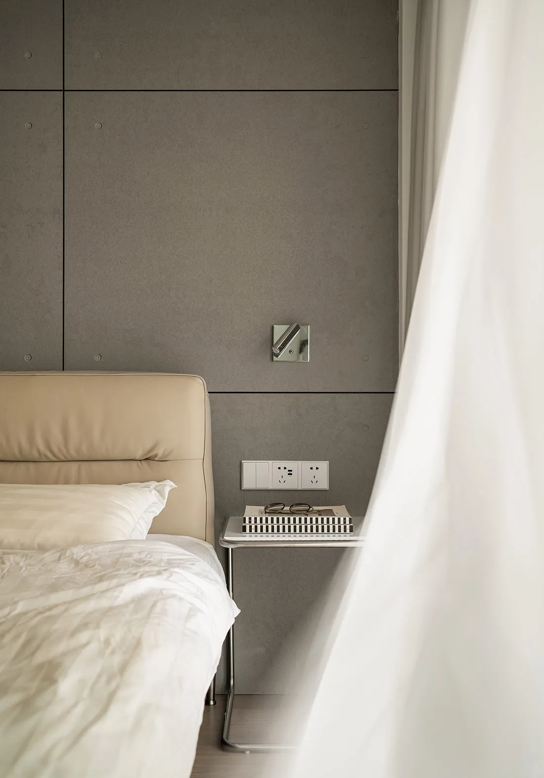 A minimalist bedroom featuring a bed with a beige leather headboard and a nightstand with a book, glasses, and power outlets