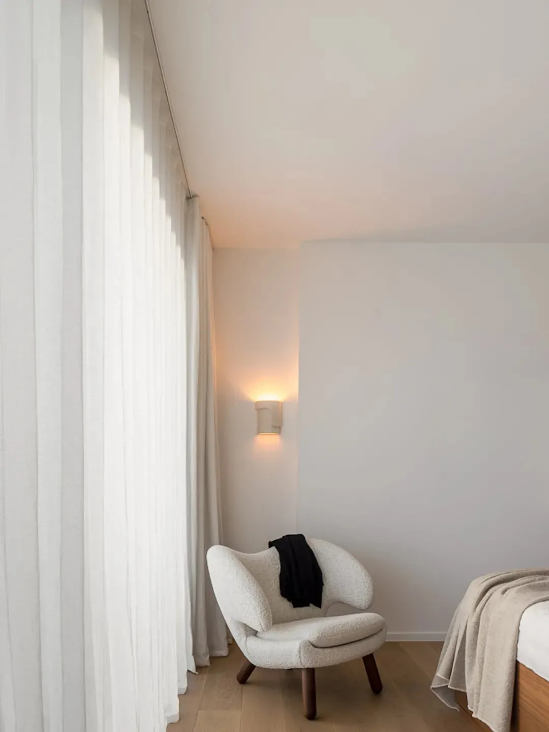 A minimalist bedroom featuring a modern upholstered chair, a wall-mounted lamp, and a wooden bed frame with neutral textiles.