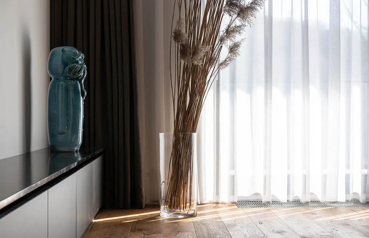 A tall cylindrical glass vase sitting on the floor between curtains and a window, containing dried grasses.