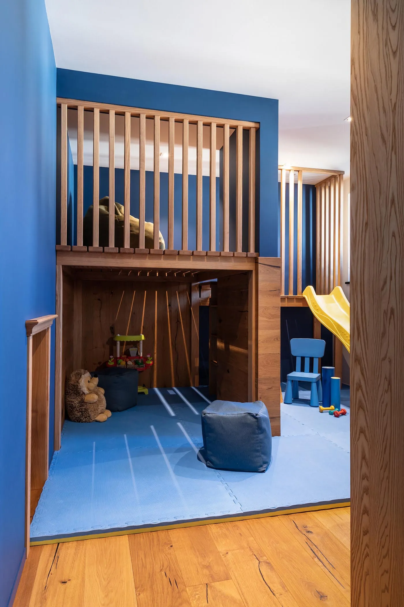 A modern playroom with wooden floors and blue walls, featuring a lofted area with a slide and various toys.