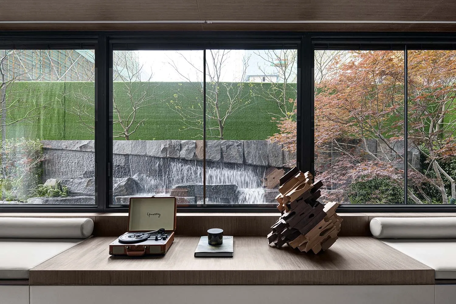 A minimalist living room with wooden furnishings and stone accents, featuring a small table with a record player, books, and
