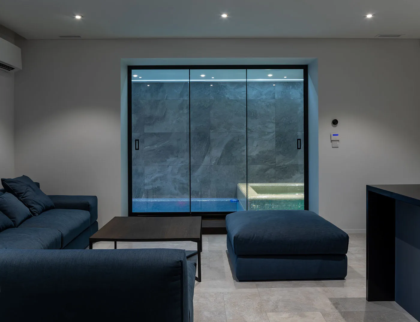 A modern minimalist living room featuring glass walls, furniture, and a blue ottoman.
