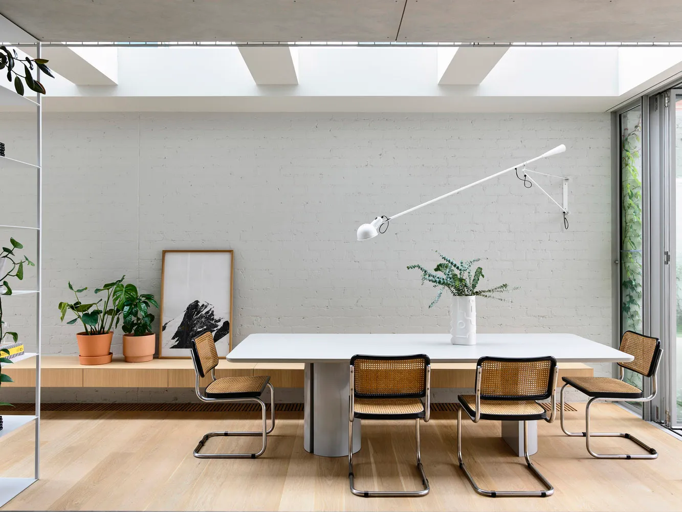 A minimalist dining room with a white table, cane chairs, and a white wall with skylights.